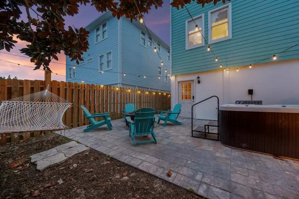 Backyard at dusk with the hot tub, hammock, and the full three-story teal exterior of the house lit up behind the patio