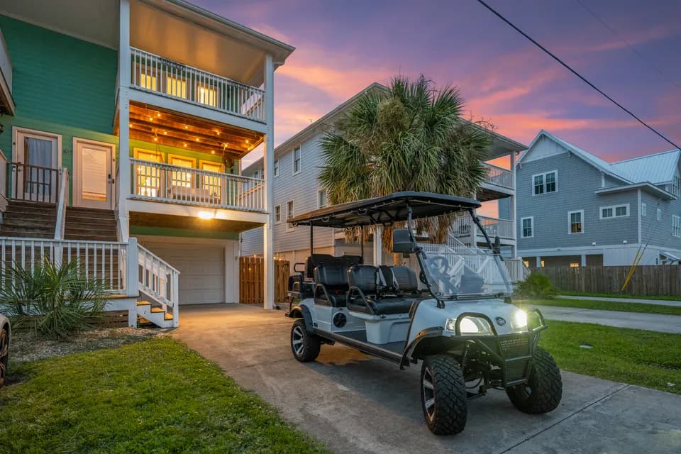 Three-story teal beach house exterior at twilight with a golf cart parked in the driveway and string lights glowing along the porch
