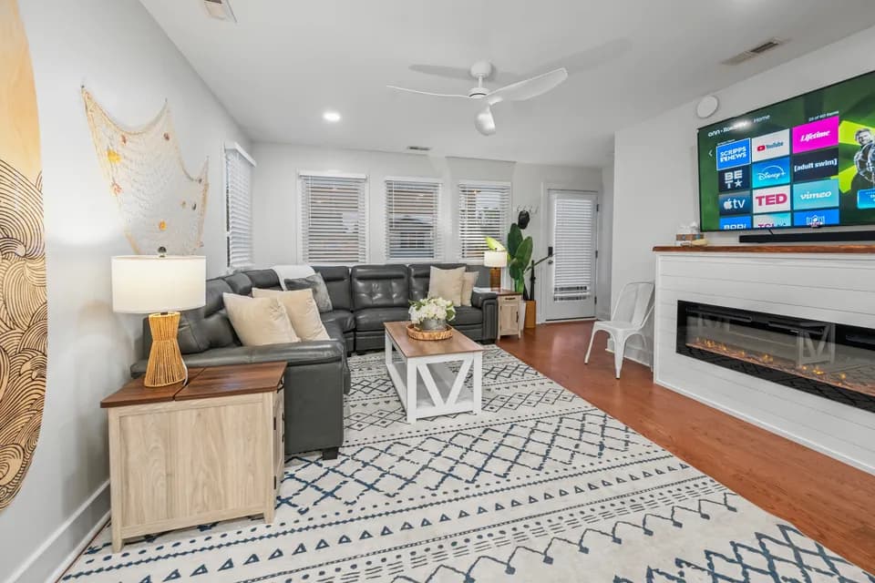 Living room with a dark gray leather sectional, blue-and-white patterned area rug, white coffee table, white built-in fireplace with mounted TV, and a surfboard and woven net wall display above the couch