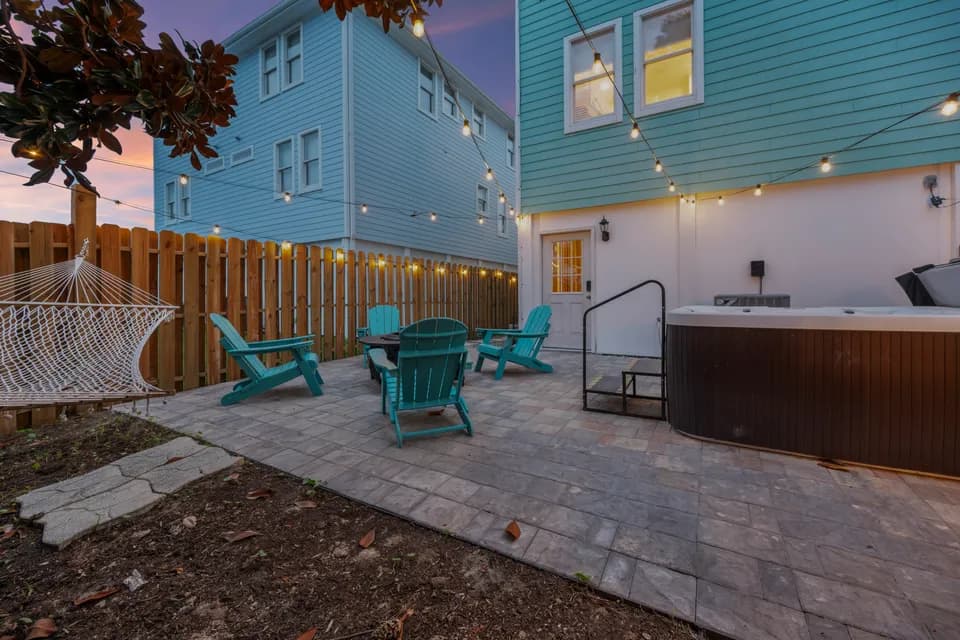 Fire pit surrounded by teal Adirondack chairs at twilight, string lights strung through the magnolia tree, wood-fenced backyard with the house glowing in the background