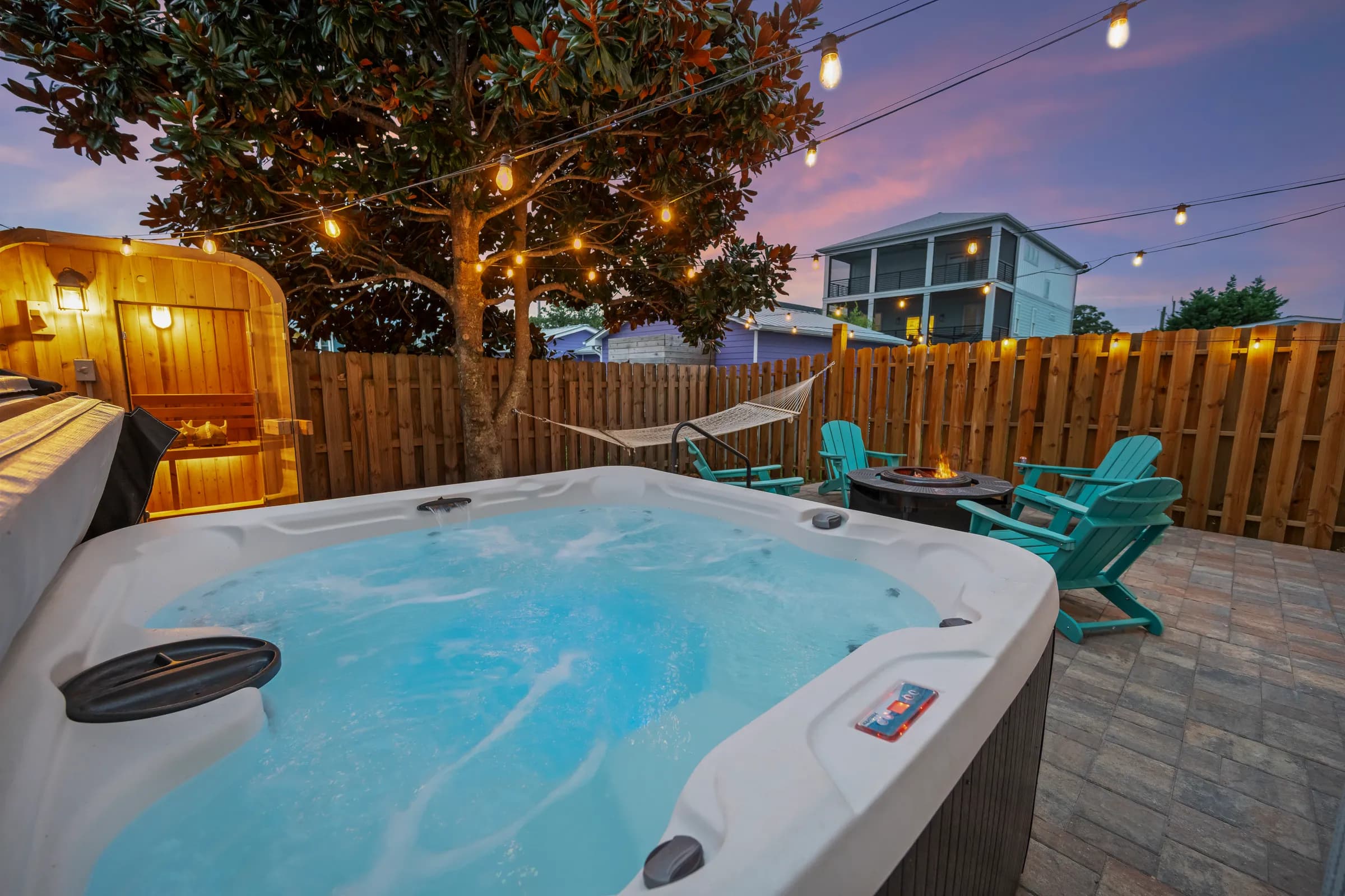 Private hot tub glowing blue at dusk beneath the magnolia tree, string lights overhead and a wood privacy fence behind it