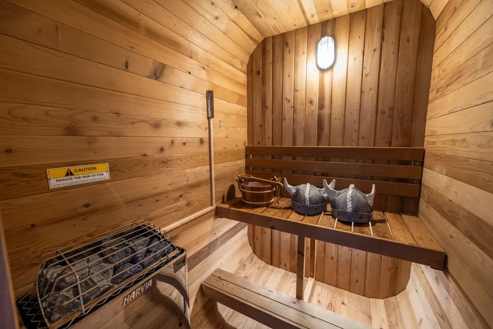 Inside the cedarwood sauna: wood-paneled walls, a warm wall sconce, and a two-person bench with a bucket and sauna hats