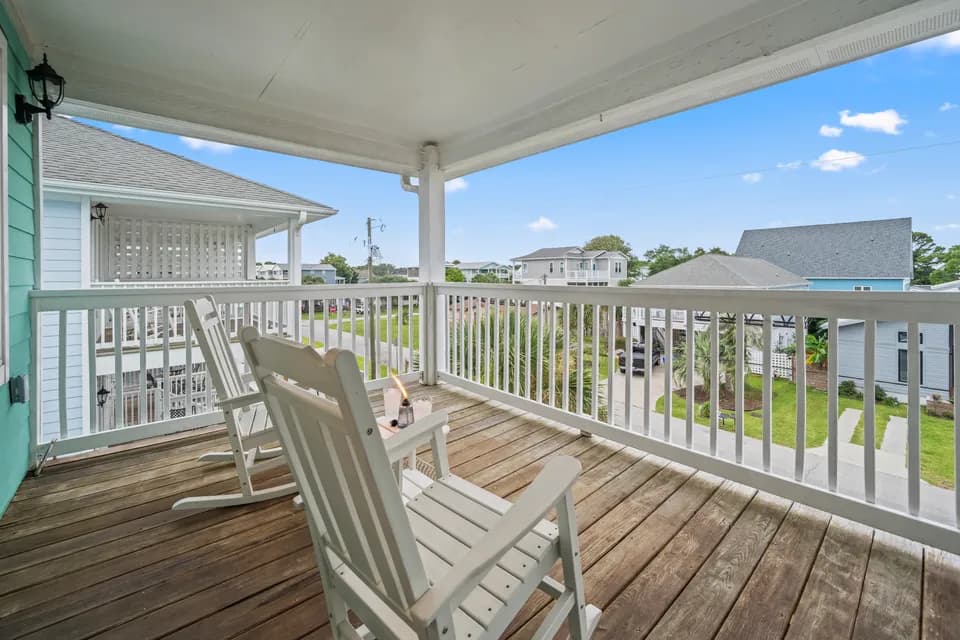 Covered upper balcony with white rocking chairs overlooking the neighborhood rooftops under a clear blue sky